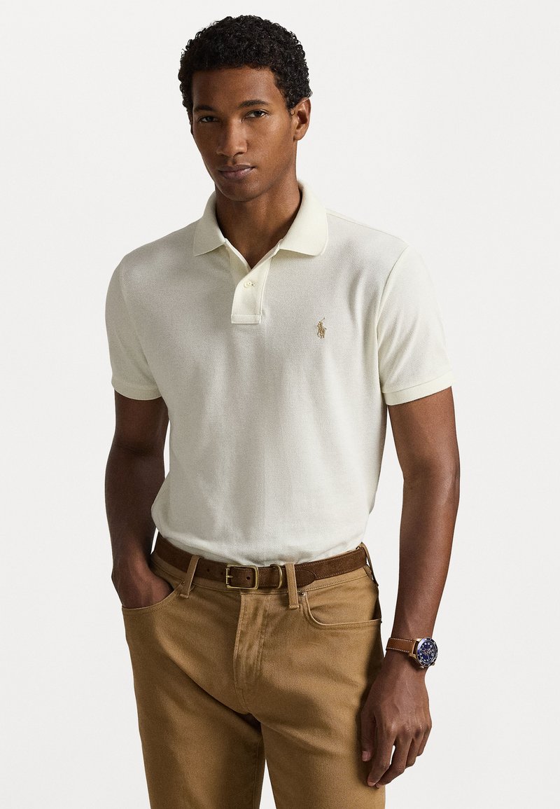 Gai dzeltenas polo krekls ar īsām piedurknēm, kas raksturojas ar smalku tekstūru un mazu izšūtu logo, kombinēts ar brūnām biksēm un pulksteni.