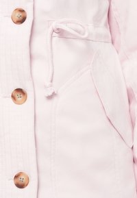 Casaco rosa claro com botões castanhos, cordão ajustável, costura detalhada e um bolso frontal em tecido suave.