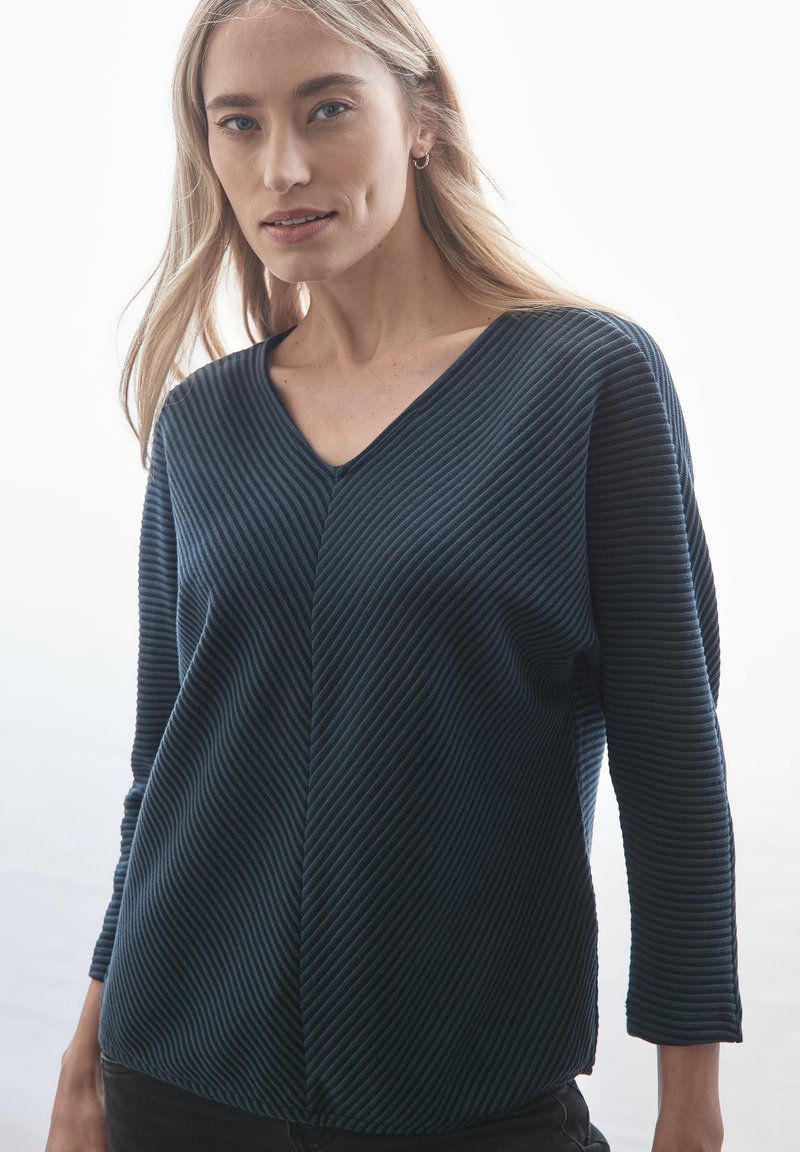 Laadukkaasti valmistettu tummansininen raitapaita pitkillä hihoilla, jossa on V-pääntie ja epäsymmetriset saumat. Pehmeä rakenne vaakasuorilla kuvioilla, hieman väljä istuvuus.