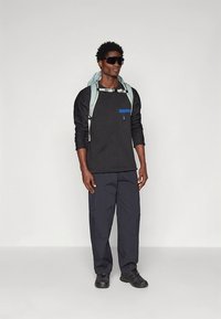 Črna majica z dolgimi rokavi s modrim žepom, kombinirana s temnimi hlačami in črnimi supergami. Nošen je svetlo moder nahrbtnik, dopolnjen z sončnimi očali.