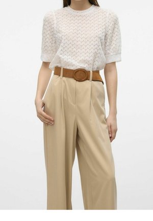 Frau trägt eine weiße Spitzenbluse mit kurzen Ärmeln, eingesteckt in beige, hoch taillierte weite Hosen mit einem braunen Wildledergürtel.