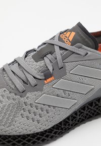 Szare buty sportowe z teksturowaną siateczką, pomarańczowymi akcentami i trzema metalicznymi, szarymi paskami. Posiadają czarną, teksturowaną podeszwę.