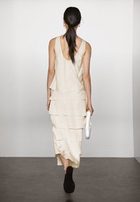 Leinenkleid mit gestuftem Design in Elfenbein, das einen tiefen Rückenausschnitt aufweist. Das Model hält eine strukturierte weiße Handtasche und trägt schwarze Schuhe.