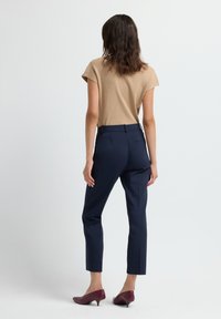 Kvinna som står vänd bort med en beige kortärmad topp, marinblå skräddarsydda byxor och bourgogne lågklackade skor på en vit bakgrund.