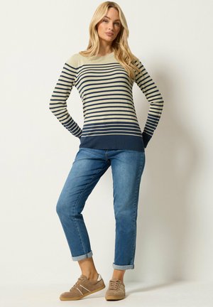 Woman wearing a beige and navy striped sweater, rolled-up blue jeans, and light brown sneakers standing against a white background.