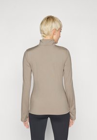 Un haut à manches longues beige avec un col montant, au design ajusté et à la texture lisse, associé à des leggings noirs, vu de dos.