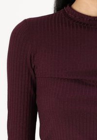 Burgundska rebrasta majica s visokim ovratnikom i prilagođenim dizajnom. Sadrži teksturirani uzorak s vertikalnim prugama i suptilnom nabranom na ovratniku.
