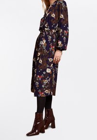 Blommönstrad klänning med lång ärm, åtdragen midja och knälång fåll i marinblått, tillsammans med glänsande bruna ankelskor.