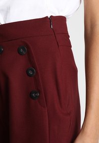 Pantalons taille haute bordeaux avec une fermeture éclair sur le côté, ornés de quatre boutons décoratifs noir et d'un tissu lisse et texturé.