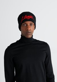 Gorro de punto negro con el logotipo "Hugo" bordado en rojo, combinado con un suéter de cuello alto negro. La tela parece suave y elástica.