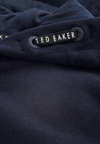 Marinblått textil med en mjuk struktur. En svart tygetikett med vit text står det "TED BAKER" på, fäst med ett runt ögla.