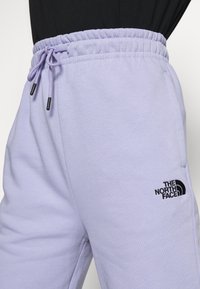 Fialové tepláky vyrobené z měkké tkaniny, s elastickým pasem na šňůrku a černým logem na levém boku.