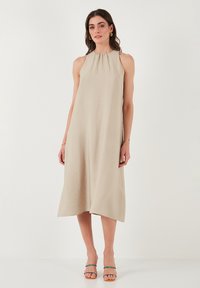 Beige A-line dress with a high neckline, sleeveless design, and textured fabric. Falls just above the ankles, paired with strappy sandals.