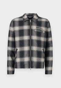 Plaid zip-up jacket in navy and cream, featuring a collar, side pockets, and a chest pocket with embroidered detail. Textured wool blend.