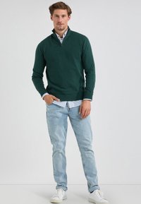 Groene pullover met een kwart-rits kraag, gelaagd over een lichtblauw gestreept shirt, gecombineerd met lichte denim jeans en witte sneakers.