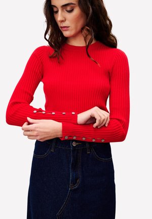 Rode geribbelde longsleeve trui met een aansluitend ontwerp. Zilveren knoopaccenten op de manchetten, gecombineerd met een donkerdenim rok.