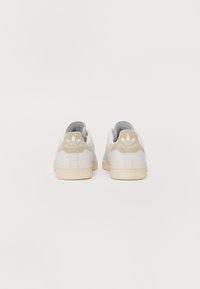 Sapatilhas brancas de couro com um tab de calcanhar em camurça bege, apresentando um detalhe de logótipo em relevo e um design simples e aerodinâmico.