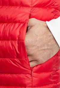 Gilet imbottito rosso con texture trapuntata, dotato di tasca con zip e mano parzialmente infilata all'interno. Cuffia bianca visibile. Finitura liscia.