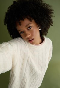 Vit kabelstickad tröja med ett strukturerat mönster och ribbad halsringning, i kombination med en ljusrosa topp under. Bakgrunden är i dämpat grönt.