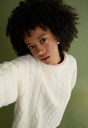 Pulover alb tricotat, cu un model texturat și guler din jerseu, asortat cu un top roz deschis dedesubt. Fundal de un verde discret.