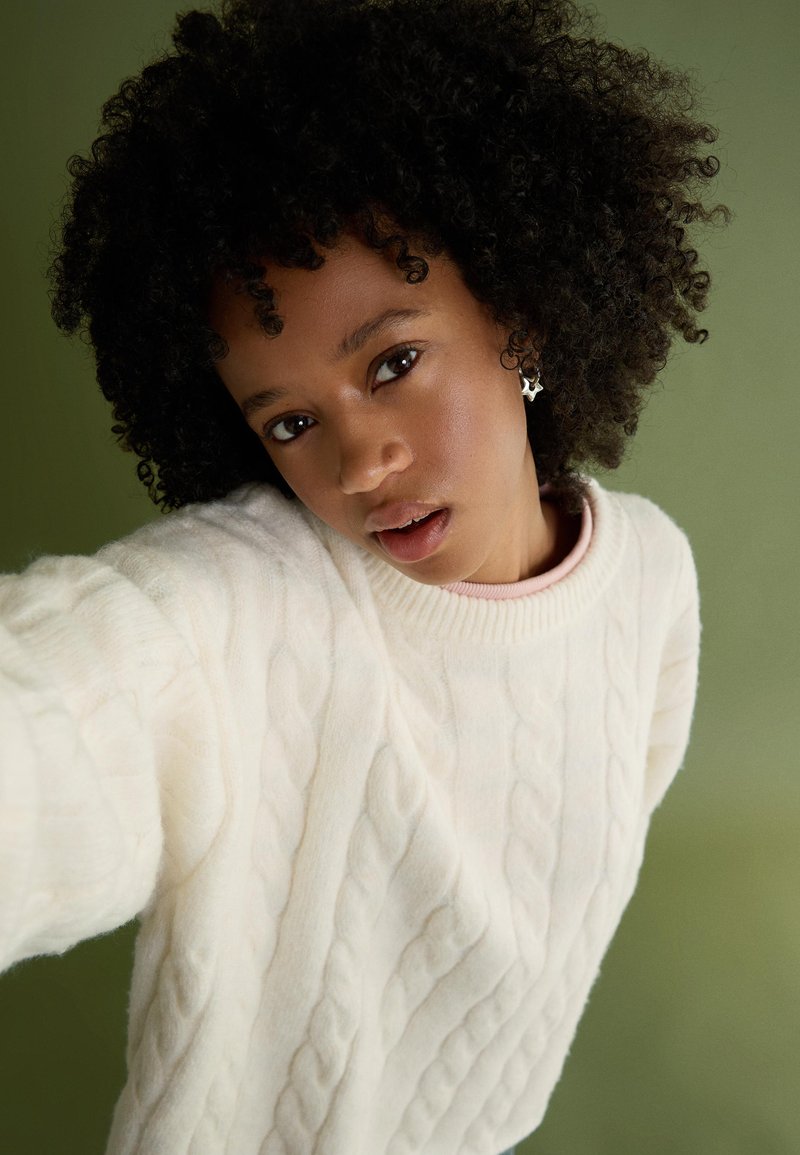 Vit kabelstickad tröja med ett strukturerat mönster och ribbad halsringning, i kombination med en ljusrosa topp under. Bakgrunden är i dämpat grönt.