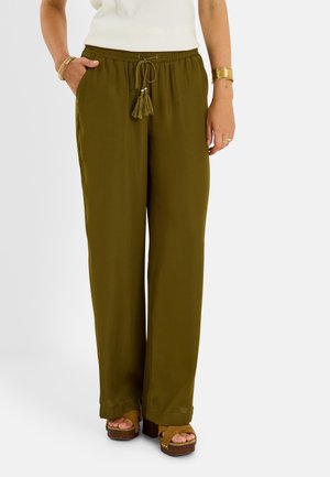 Mujer con pantalones anchos de color verde oliva con cordón de borlas, una camiseta blanca, pulseras doradas, anillos y sandalias de plataforma marrones.