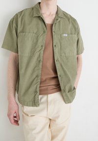 Camicia verde oliva a maniche corte con due tasche frontali, chiusura a bottoni, indossata sopra una canotta marrone e pantaloni chiari.