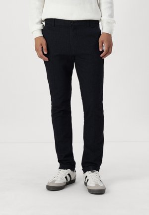 Personne portant un pantalon slim noir, des baskets blanches et noires, et un pull blanc côtelé, debout devant un fond clair uni.