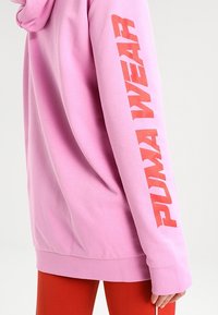 Felpa rosa con una vestibilità comoda, che presenta un grande logo "PUMA WEAR" arancione sulla manica sinistra. Tessuto morbido e tasca a marsupio.