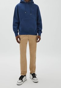 Blauer Kapuzenpullover aus Baumwolle mit Fronttasche und Kordelzug, kombiniert mit hellbraunen Chinos und weißen Sneakers mit schwarzen Akzenten.
