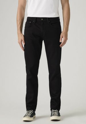 Mężczyzna ubrany w czarne obcisłe dżinsy, białą koszulkę i czarne trampki z białymi sznurowadłami, stojący na jednolitym tle.