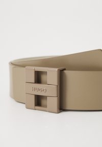 Beige műbőr öv, négyzet alakú fém csattal, amelyen a "HUGO" felirat látható. Sima tapintás és letisztult dizájn jellemzi.