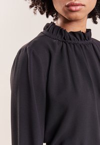 Donna che indossa una blusa nera a maniche lunghe con un colletto alto arricciato, ritagliata dal naso fino a metà torso.