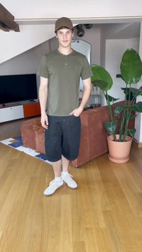 Camisa henley verde de mangas curtas, calções de ganga pretos, ténis brancos. Ambiente interno com plantas de interior e um sofá castanho ao fundo.