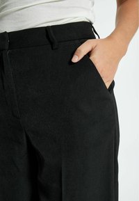 Černé kalhoty s hladkou texturou, vybavené bočními kapsami, těsným pasem a zapínáním na zip. Jednoduchý a moderní design.