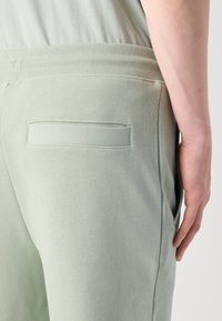 Pantalones de chándal verde claro con una cinturilla acanalada, que presentan un pequeño bolsillo trasero y una tela suave y texturizada. Detalle del lado de la prenda.