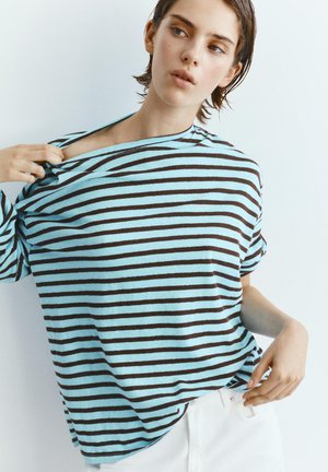 Tânăr care își ajustează o cămașă în dungi bleu deschis și negru, purtând pantaloni albi, pe un fundal simplu și deschis la culoare.