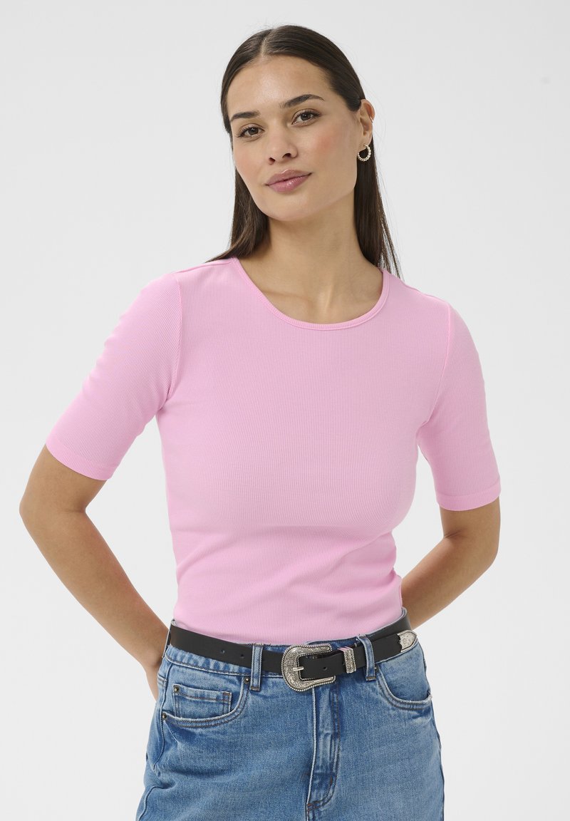 Žena s dlhými tmavými vlasmi, oblečená v ružovom rebrovanom tričku s krátkym rukávom, modrých džínsoch, čiernom opasku so striebornou prackou a kruhových náušniciach, stojaca s rukami za chrbtom.