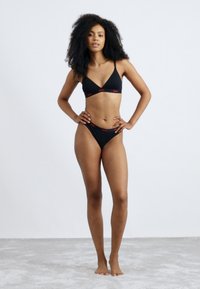 Zwarte driehoekige bikini-top met verstelbare bandjes en bijpassende bikini-bottom. Beide hebben rode logo-accenten. Glad, rekbaar materiaal.