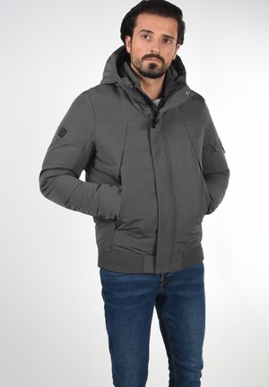Veste grise matelassée avec capuche, dotée d'une fermeture éclair sur le devant, de poches latérales, de poignets ribbed et d'une texture extérieure lisse. Portée avec un jean bleu.