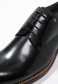 Chaussure de ville en cuir verni noir avec lacets fermés, couture en cuir marron, et bout arrondi lisse, présentée en gros plan.