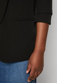 Black blazer with a folded cuff and a side pocket, paired with blue denim jeans. Smooth texture and tailored design.
