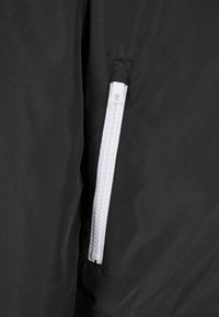 Svart jacktyg med vit dragkedja på sidan, som framhäver den släta strukturen och den minimalistiska designen.