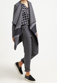 Grijze, gelaagde open cardigan met zwarte accenten, gecombineerd met een marineblauw geblokt shirt en grijze taps toelopende broek. Zwarte instap schoenen maken de outfit compleet.