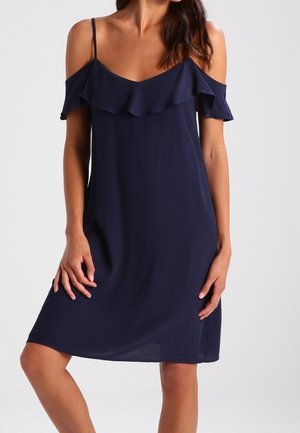 Femme portant une robe bleu marine épaules dénudées avec fines bretelles et un col à volants, debout devant un fond clair uni.