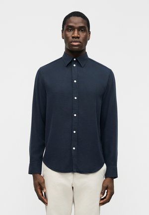 Μαύρος άνδρας που φοράει σκούρο μπλε πουκάμισο με λευκά κουμπιά και ανοιχτό μπεζ παντελόνι, μπροστά σε απλό φωτεινό φόντο.