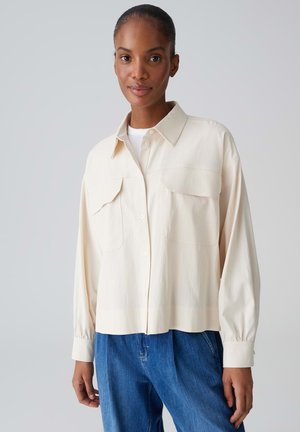 Frau mit zurückgebundenem Haar, die ein cremefarbenes Hemd mit Knöpfen und Brusttaschen sowie blaue Jeans trägt, stehend vor einem schlichten hellen Hintergrund.