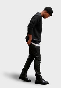 Felpa nera con texture, pantaloni neri aderenti e stivali neri lucidi con lacci. L'outfit ha un design minimalista. Il modello è in posizione laterale.
