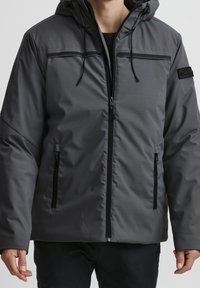 Grå isolerad jacka med huva, dragkedja fram och två sidofickor med svarta accenter samt en slät, vattenavvisande ytfinish.