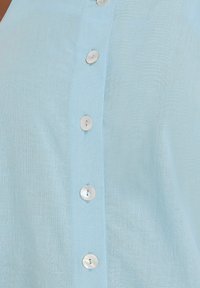 Camicia di lino azzurro chiaro con un design a bottoni sul davanti, caratterizzata da bottoni circolari bianchi e una finitura in tessuto testurizzato.